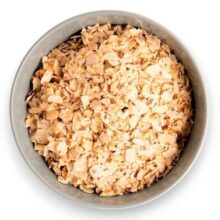 Bowl of light brown toasted oat flakes (granola) in a round metal dish, viewed from above.